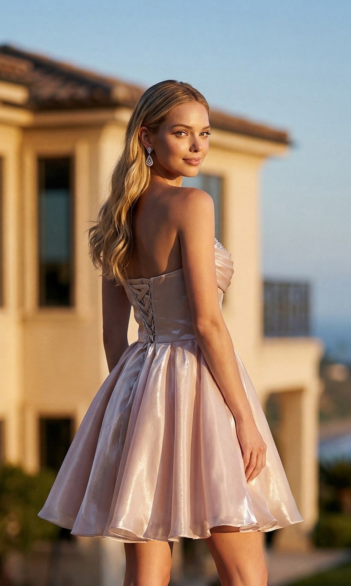 A woman wearing the Short Homecoming Dress: Juliet JT7187S—an elegant strapless dress crafted from shimmer organza with a corset bodice and lace-up back—stands outside near a beige building, glancing over her shoulder.